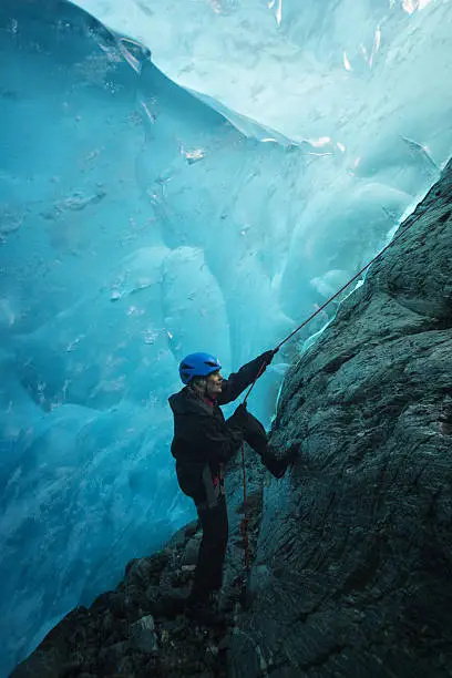 Blond woman rappells down granite bedrock into a blue ice cave and reaches a rocky ledge within Lemon Glacier, Juneau Icefield, Juneau, Alaska, USA
