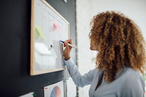 Side view of latin businesswoman standing near whiteboard and writing. She is making schedule for next business week.