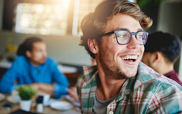Sharing laughs over coffee Shot of a smiling young man sitting at a cafe table with friends in the background group of people wearing eyeglasses stock pictures, royalty-free photos & images