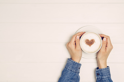 Female hand holding cup of coffee on white wooden table. Photograph taken from above, top view with copy space