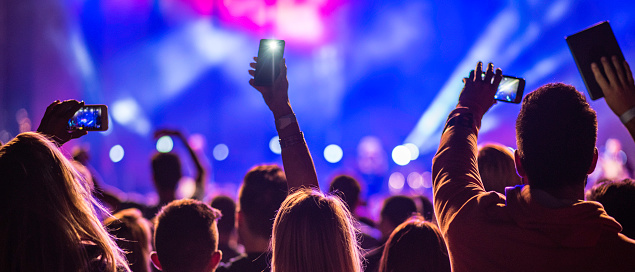 Rear close up view of a few people at a concert holding their smartphones above their heads and filming the performance.