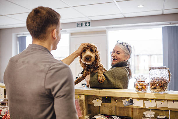 Time For a Haircut Woman is holding her dog at the reception counter of the grooming salon. She is booking her dog in to be groomed. There is a man working on reception and he is stroking the dog. pet shop photos stock pictures, royalty-free photos & images