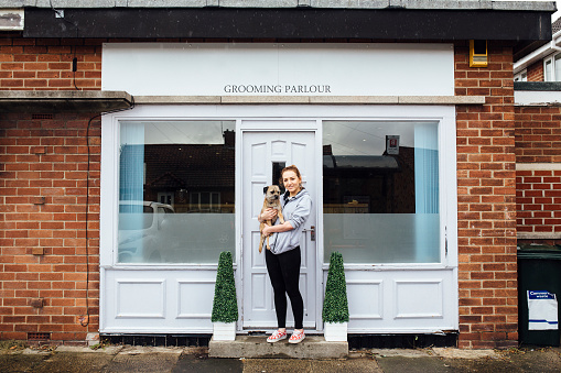 Young business-owner is posing for the camera outside of her grooming parlour with her pet dog in her arms.