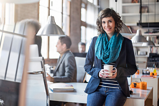Portrait of confident mid adult businesswoman holding coffee cup. Smiling female professional is leaning on table. She is with colleagues in creative office.