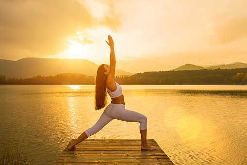 Woman doing a warrior I yoga pose at sunset over lake