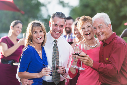 A group of people socializing at an outdoor party. The focus is on the two couples in the foreground standing together, smiling at the camera. The senior couple on the right holding wine glasses are in their 70s.