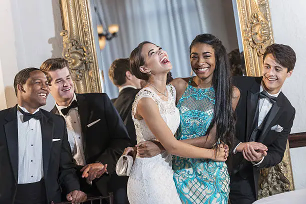 A group of five multi-ethnic teenagers and  young adults dressed in formalwear - dresses and tuxedos. They are at a special event, perhaps a prom. The two girls are standing in the middle, laughing and hugging at the men watch and applaud.