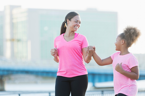 A black mother and daughter walking together outdoors along the waterfront of a city. They are holding hands, making eye contact and smiling at each other lovingly. They are pink shirts.