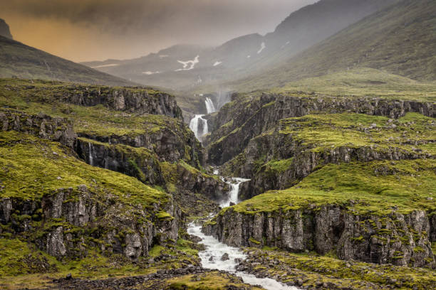 Klifbrekkufossar waterfalls situated in the bottom of the Mjóifjörður fjord. The waterfalls are in the river Fjarðará and cascade down in a stair-step pattern amidst grassy slopes sheltered by murky cliff walls.