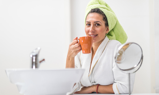 Woman wearing bathrobe and sitting in the bathroom drinking coffee in the morning