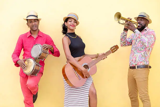 Cuban musical band, the trio consisting of a well known musicians standing against the bright yellow wall. Beautiful young woman standing in the middle, holding a guitar. The man on the left holding the small drums bongos, and a musician on the left holding a trumpet. Havana, Cuba, 50 megapixel image.