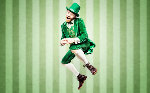 A stereotypical Irish character all ready for Saint Patricks day.  He dances, jumping high in the air during a jig with a big smile on his face.  Studio portrait.  Horizontal image with copy space.  Green striped wallpaper background.