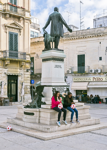 Lecce. Italy - December 22. 2016: Two adolescent females holding footballs engrossed in electronic devices, seated on the Sigismondo Castromediano monument in the Piazzetta after his name, in Lecce, Italy.