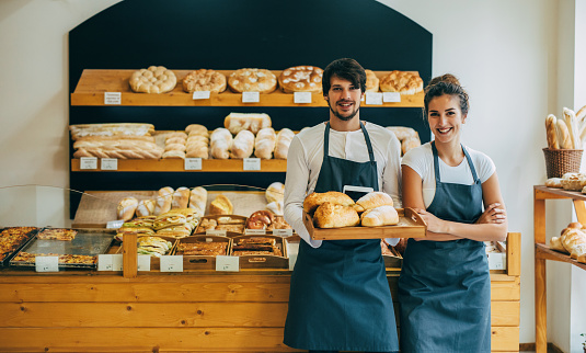 Two beautiful young bakers holding a tray of bread at the bakery.