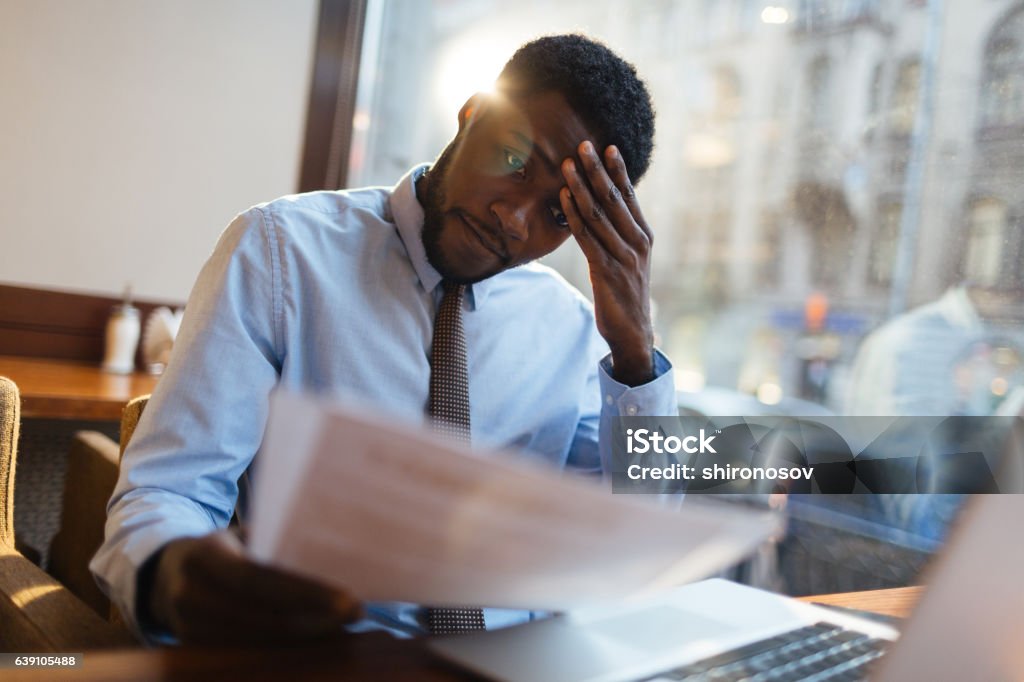 Confused manager in cafe Focused African-American office manager sitting at cafe table with laptop, reading important documents with puzzled expression and holding head with his hand. Emotional Stress Stock Photo