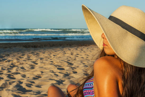 Girl sitting on the beach wearing beach summer hat turned stock photo