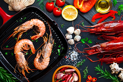 Top view of an iron grill with three shrimps on a kitchen counter top. The grill is at the left of the frame while some ingredients for cooking shrimps like olive oil, salt and pepper, garlic, lemon and herbs are at the right. DSRL studio photo taken with Canon EOS 5D Mk II and Canon EF 100mm f/2.8L Macro IS USM