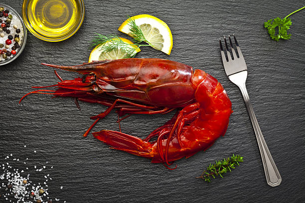 Top view of a single giant red shrimp shot on dark table ready to eat. A fork is at the right side of the crustacean. Some ingredients for preparing shrimp like olive oil, salt and pepper, lemon and herbs are all around the shrimp. DSRL studio photo taken with Canon EOS 5D Mk II and Canon EF 100mm f/2.8L Macro IS USM