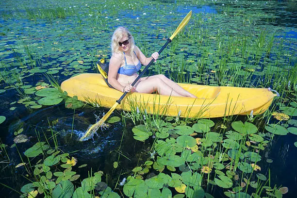 Caucasian young woman in her 20's wearing a blue bikini  paddles a yellow kayak through lilypads on a summer day, Ten Mile Lake, Walker, Minnesota, USA
