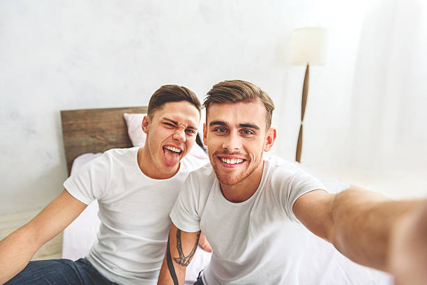 Cheerful homosexual couple making selfie in bedroom Joyful two gays are having fun at home. One man is stretching arm and photographing them. They are sitting on bed and smiling gay man photos stock pictures, royalty-free photos & images