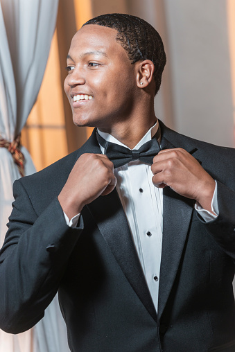Young black Hispanic man wearing a tuxedo. He is smiling, dressed for a special event, perhaps a wedding or prom. He is adjusting his bowtie.