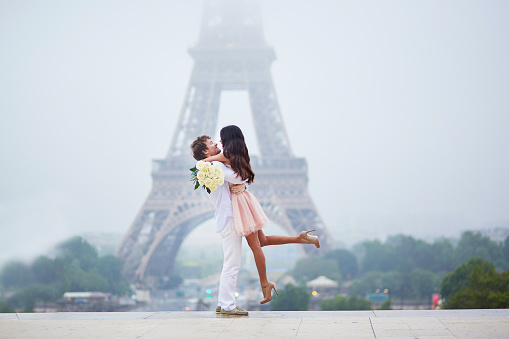 Beautiful romantic couple in love with bunch of white roses having fun near the Eiffel tower in Paris on a cloudy and foggy rainy day