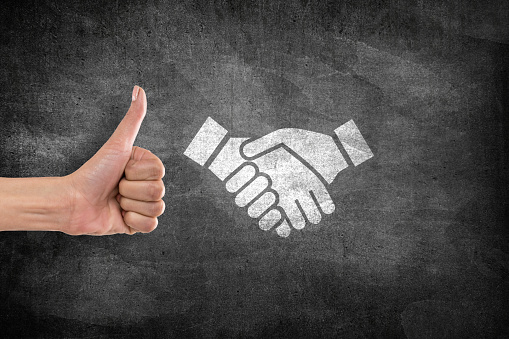Businesswoman agrees to handshake icon with thumbs up gesture in front of blackboard, successful business deal