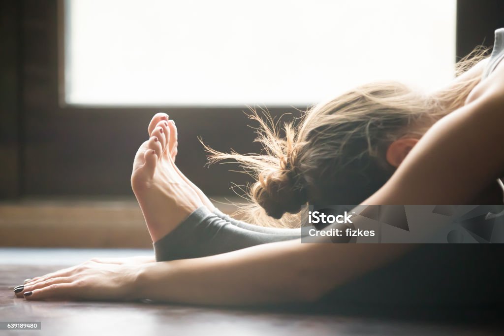Young woman in paschimottanasana pose, home interior background, Young woman practicing yoga, sitting in Seated forward bend exercise, paschimottanasana pose, working out, wearing sportswear, grey pants, bra, indoor, home interior background, close up Yoga Stock Photo