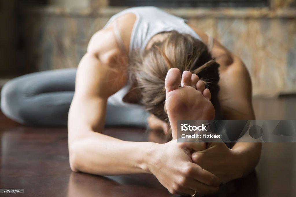 Close up young woman in Janu Sirsasana pose, home interior Close up of young woman practicing yoga, sitting in Head to Knee Forward Bend exercise, Janu Sirsasana pose, working out, wearing sportswear, grey pants, bra, indoor, home interior background Yoga Stock Photo