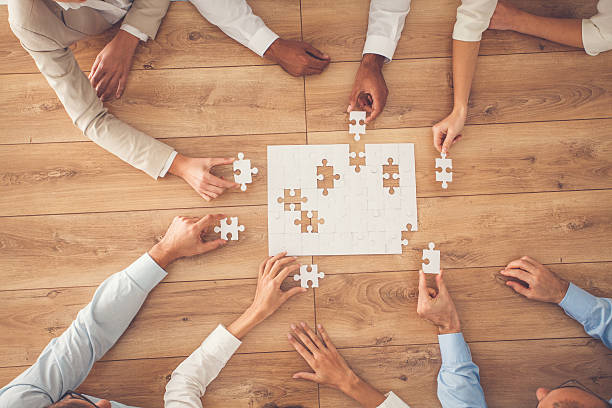 Business people finding solution together at office Business people sitting at office desk, putting puzzle pieces together, finding solution, high angle view. jigsaw piece stock pictures, royalty-free photos & images