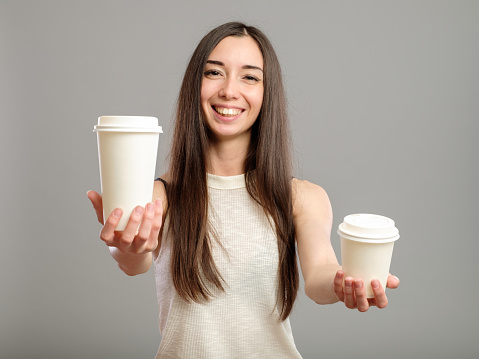 Young beautiful woman offers white cups of coffee isolated on gray background