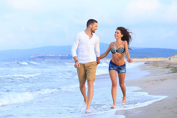 Happy young couple holding hands and walking on beach. Looking at each other. Both with casual clothes. Sea and sand on background.