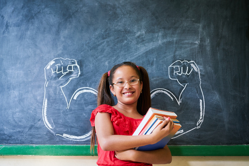 Concept on blackboard at school. Intelligent and successful hispanic girl in class. Portrait of female child smiling, looking at camera, holding books against drawing of muscles on blackboard