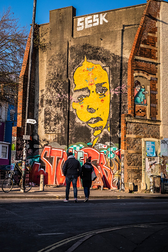 Bristol, United Kingdom - January 21, 2017: Colour image of a couple crossing a road in front of a wall with colourful grafitti and street art,  typical of that seen in the Stokes Croft area of Bristol, UK