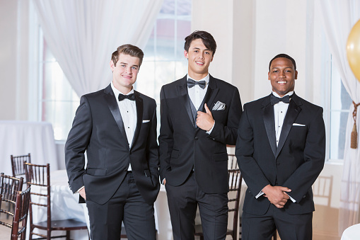 A group of three multi-ethnic young men wearing tuxedos, standing in a row, smiling at the camera. They are at a party, prom, a wedding or some other special formal event.