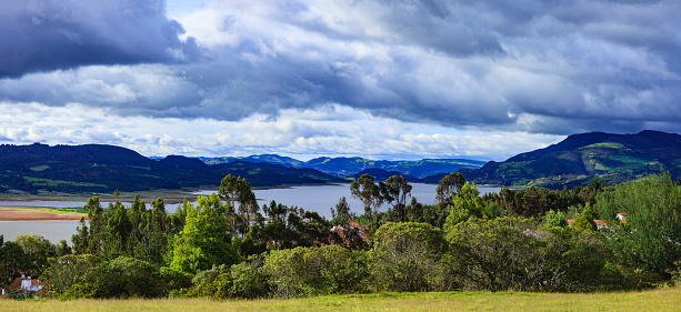 Colombia, South America - Looking across some trees towards the Andean reservoir called Embalse del Tominé in Guatavita, in the Cundinamarca department of the South American country of Colombia. The water level in the reservoir is lower than normal due to the El Niño phenomenon in early 2016. The elevation is about 8660 feet above mean sea level. In the background are the always present Andes mountains. The sky is overcast: it threatens to rain. Photo shot in the afternoon sunlight; horizontal format. Copy space. Panoramic image created by merging two images in Photoshop.