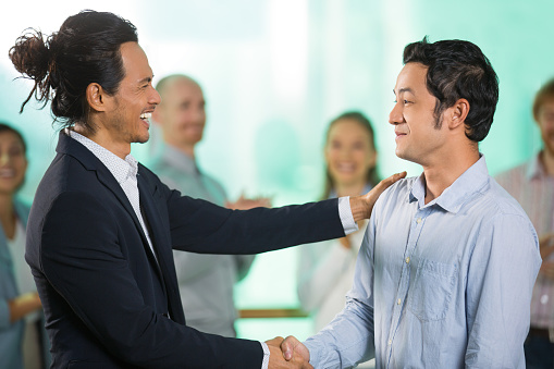 Smiling joyful middle-aged Asian businessmen shaking hands after meeting with four blurred applauding people in background. Side view.