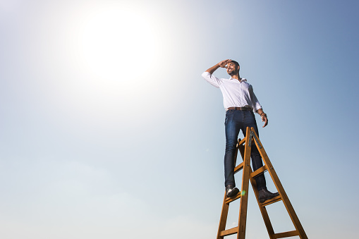 Low angle view of happy African American businessman standing on the top of the ladders and shielding his eyes while looking at distance. Copy space.