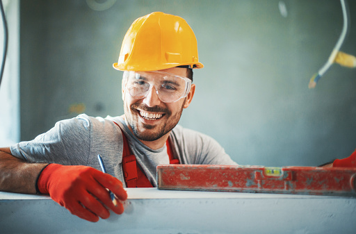 Closeup front view of a cheerful handsome construction worker leaning against a white wall and looking at camera. He's wearing red protective gloves and yellow work helmet.