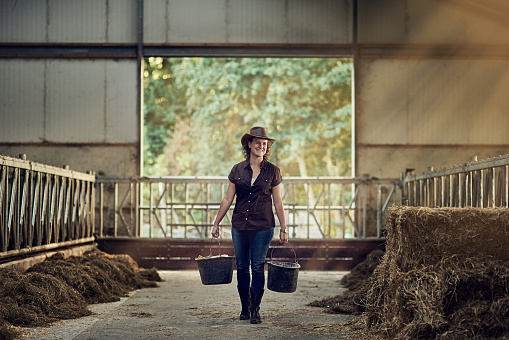 Shot of a happy female farmer carrying buckets while walking out of a barn