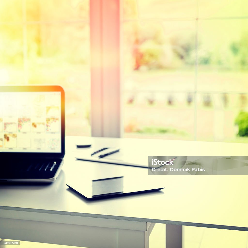 Laptop and tablet on desk in front of window, vintage New business. Laptop and tablet are standing on a table in front of a big window with garden view, home interior. Nobody at the desk. Vintage style, dusk light. Business Stock Photo