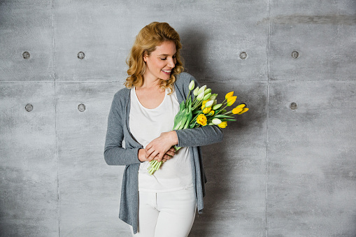 Beautiful woman holding tulips in her arms and standing against gray wall