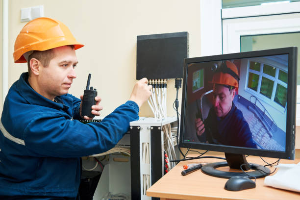 A worker wearing an orange hard hat and blue jacket is using a walkie-talkie while adjusting connections on a device, with a monitor displaying a live video feed of another person wearing a hard hat in a different location.