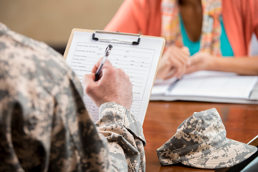 Caucasian male veteran in camouflage uniform completes paperwork for his psychiatric appointment. The female counselor is in the background.