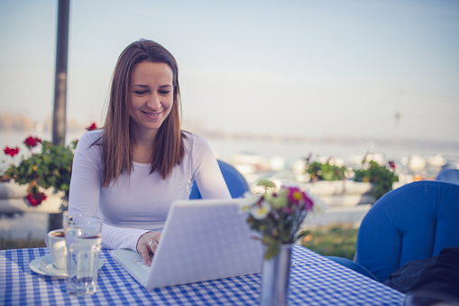 Young brunette woman enjoying the sunny morning at the beach restaurant with a cup of coffee and a laptop computer.