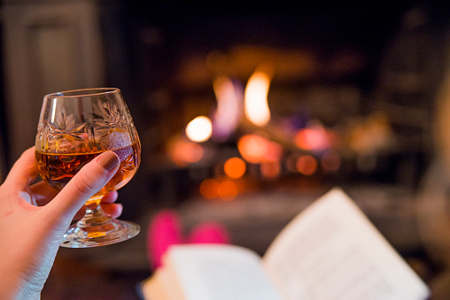 Close up of Woman resting with book near fireplace and holding glass of Whiskey