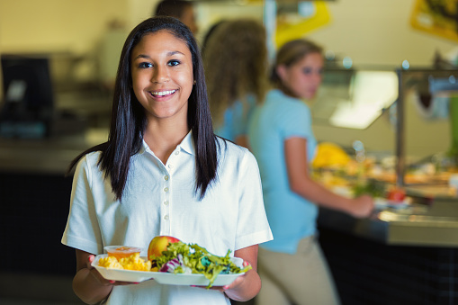 Pretty African American private high school student holds a plate of healthy food in the school cafeteria. Her classmates are waiting in line in the background. She is wearing a school uniform.