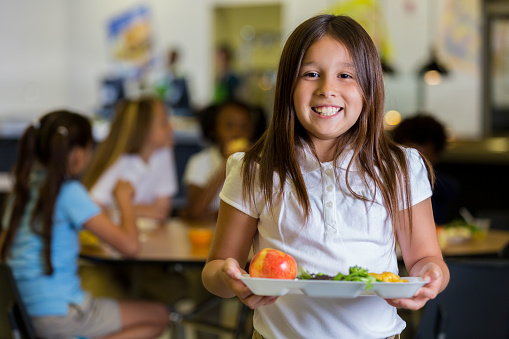 Beautiful Hispanic elementary school girl holds a plate of healthy food in the cafeteria. Her friends are eating at tables in the background. They are wearing school uniforms.