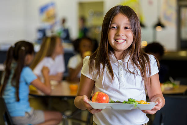 Happy elementary school girl with healthy food in cafeteria Beautiful Hispanic elementary school girl holds a plate of healthy food in the cafeteria. Her friends are eating at tables in the background. They are wearing school uniforms. tray stock pictures, royalty-free photos & images