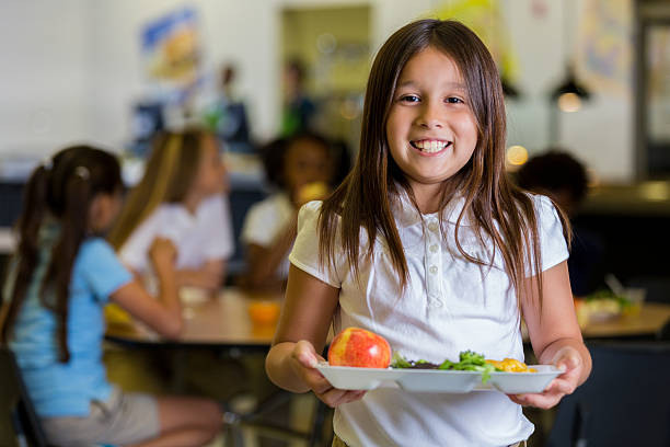 Happy elementary school girl with healthy food in cafeteria Beautiful Hispanic elementary school girl holds a plate of healthy food in the cafeteria. Her friends are eating at tables in the background. They are wearing school uniforms. school lunch child food lunch stock pictures, royalty-free photos & images